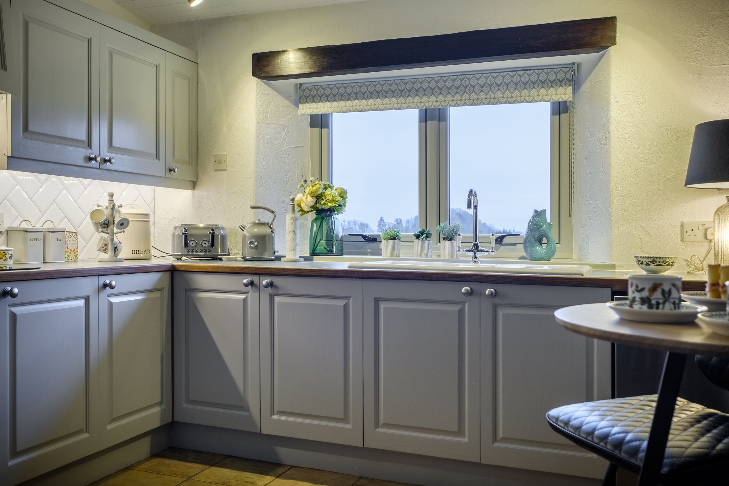 Una cocina con armarios blancos, una ventana con vistas, un lavamanos y una mesita con una silla.