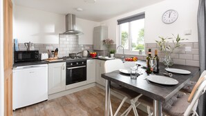 A modern kitchen with white cabinetry, a built-in oven, a microwave, and a dining area with a table set for two. - Wheal Jewel Lodge (Helston)