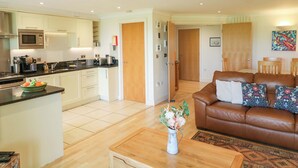 A modern kitchen with a microwave, toaster, and sink; a living area with a brown leather sofa and a coffee table; and a hallway leading to a bedroom. - 5 Thurlestone Beach (Kingsbridge)