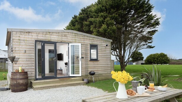A small wooden house with a sliding glass door, a barbecue outside, and a picnic table with food. - Bushwood Lodge (Helston)