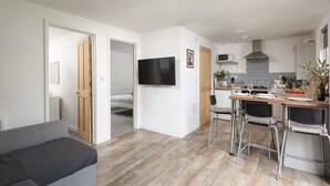 A modern apartment with a living room, a dining area, and a kitchen. There is a TV mounted on the wall, a dining table with chairs, and a kitchen counter with a microwave and stove. - Bushwood Lodge (Helston)