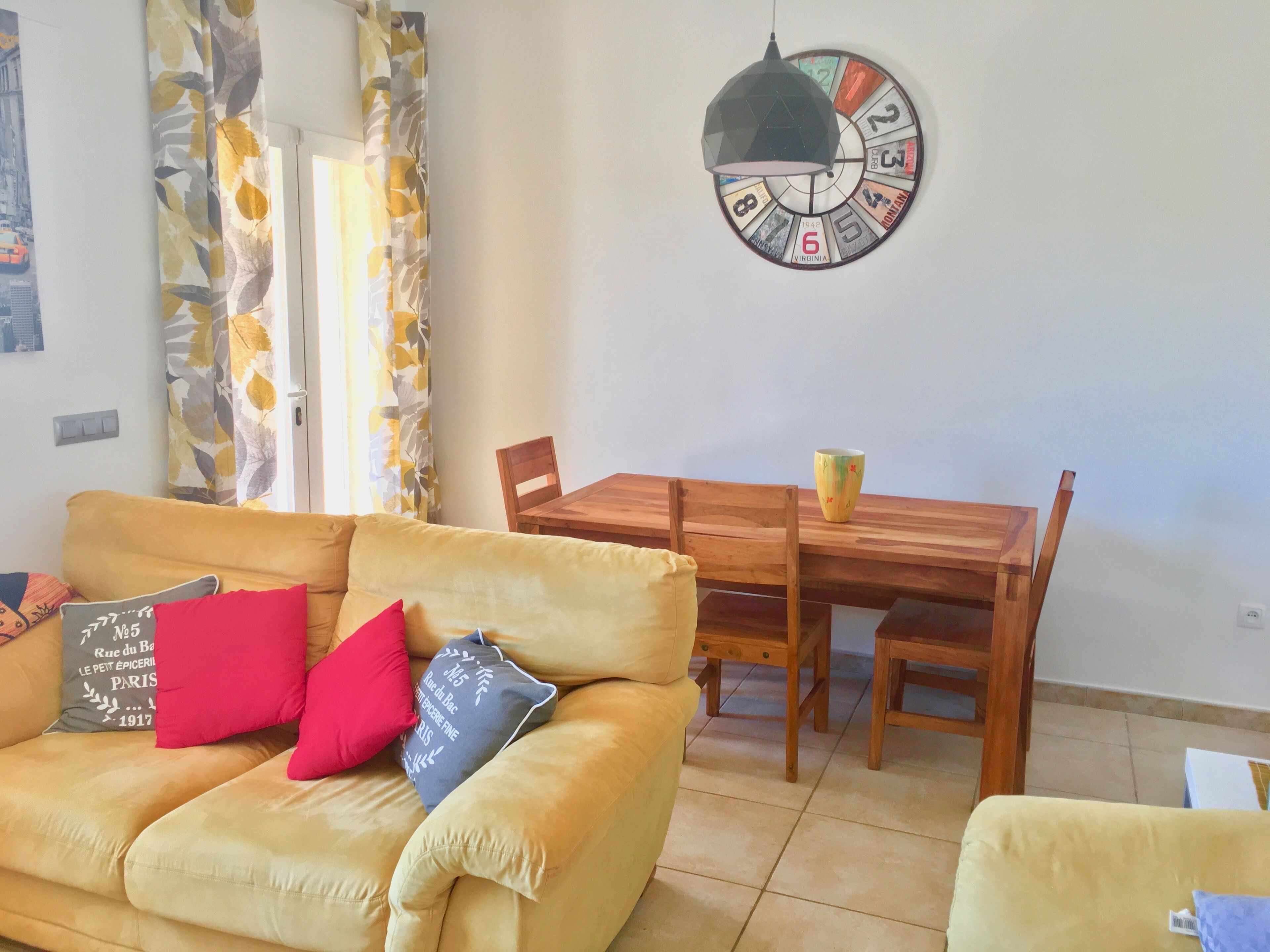 Un salon avec un canapé jaune, une table à manger en bois et une horloge murale.
