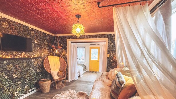 A cozy room with a patterned wall, a woven chair, a sofa with a fur throw, and a bed with a yellow door in the background. - -Shingle Diggins Cottage- (Coloma)