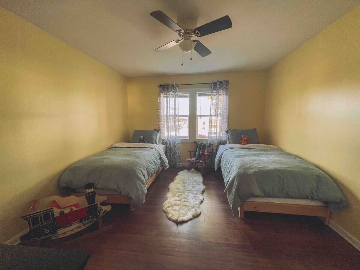 Una habitación con dos camas, un ventilador de techo, una ventana con cortinas y un tren de juguete en el suelo.