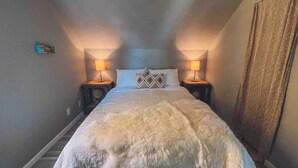 Ein Schlafzimmer mit einem großen Bett, zwei Nachttischlampen, einer flauschigen weißen Tagesdecke und einem Vorhang am Fenster.