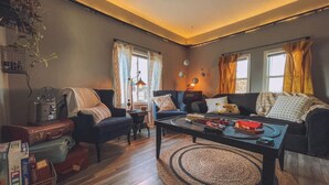 Ein gemütliches Wohnzimmer mit Sofa, Sesseln, einem Couchtisch und einem Fernseher. Vor den Fenstern hängen Vorhänge und von der Decke hängt eine Pflanze.
