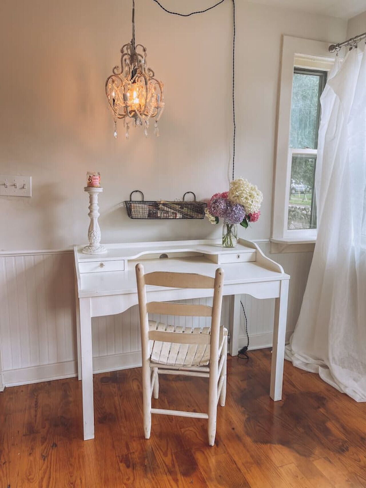 A white desk with a chair, a hanging light fixture, a vase with flowers, and a window with a curtain.
