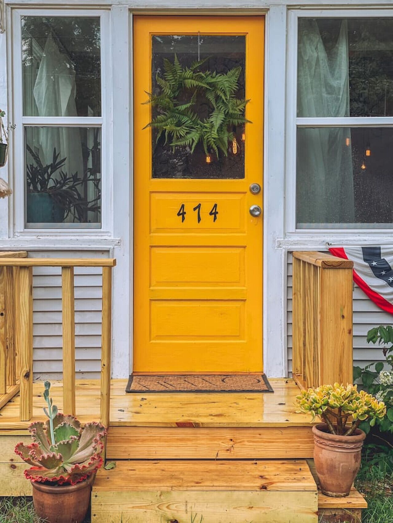 A yellow door with the number 414, a small porch with wooden railings, and two potted plants on the steps.
