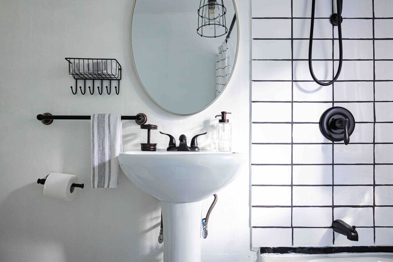 A bathroom with a white sink, a mirror, a towel rack, a toilet paper holder, and a shower with a black hose.