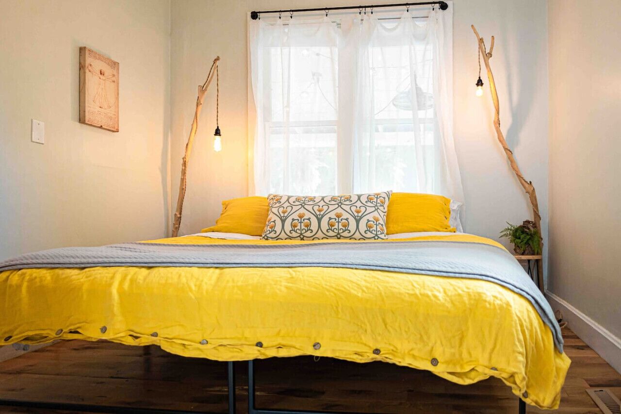 Une chambre à coucher avec un lit recouvert d’une couette jaune, un oreiller à motifs et un rideau blanc.