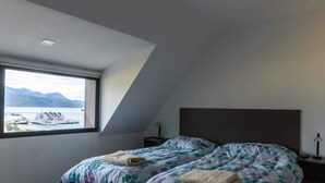 A bedroom with a bed, a nightstand, a lamp, and a window with a view of boats and mountains. - Cumbre (Ushuaia)