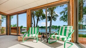 Una habitación soleada con dos sillones a rayas verdes y una mesita, ventanas amplias con vistas a un lago y árboles.