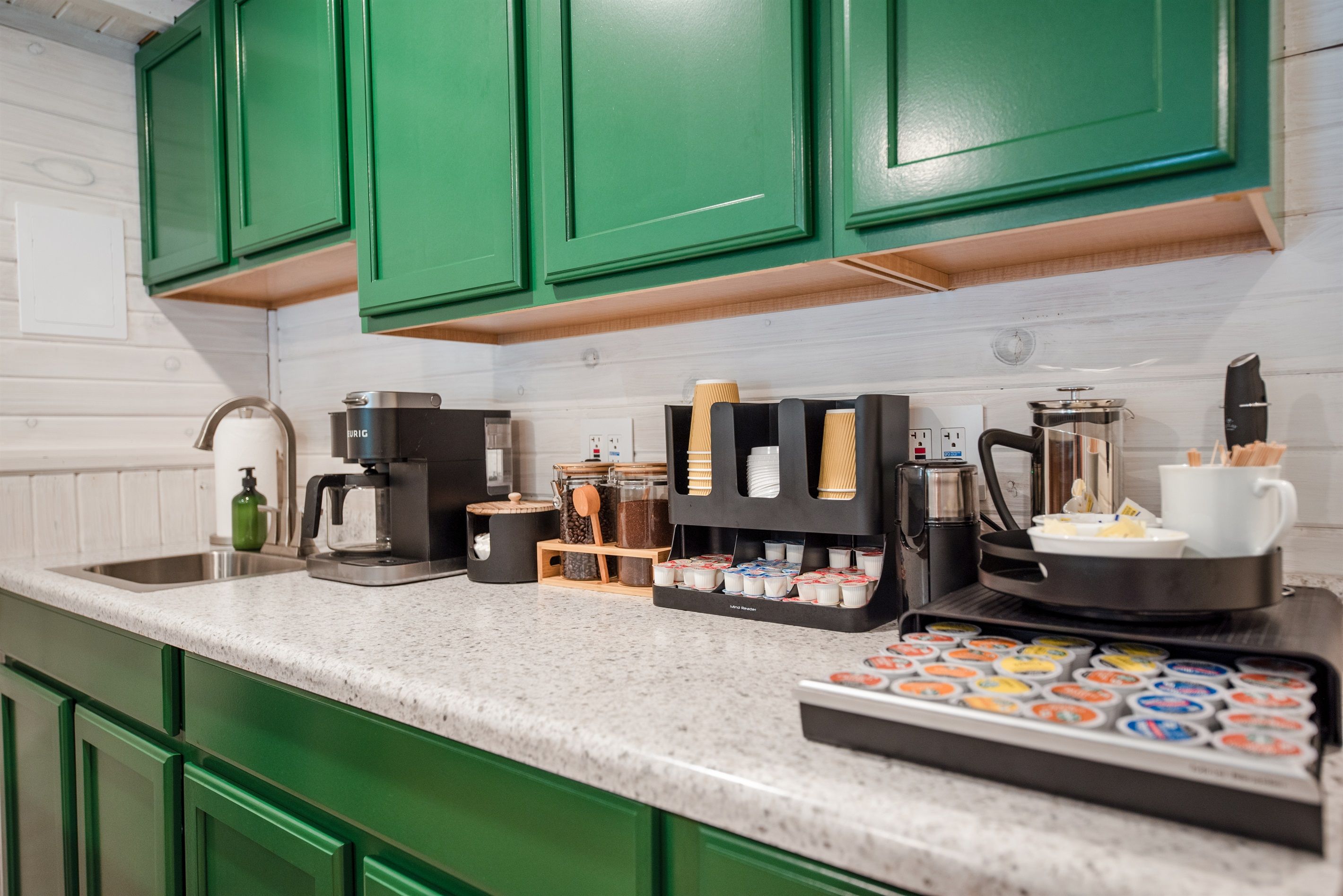 A kitchen with green cabinets, a coffee machine, a coffee press, and a tray of coffee pods.