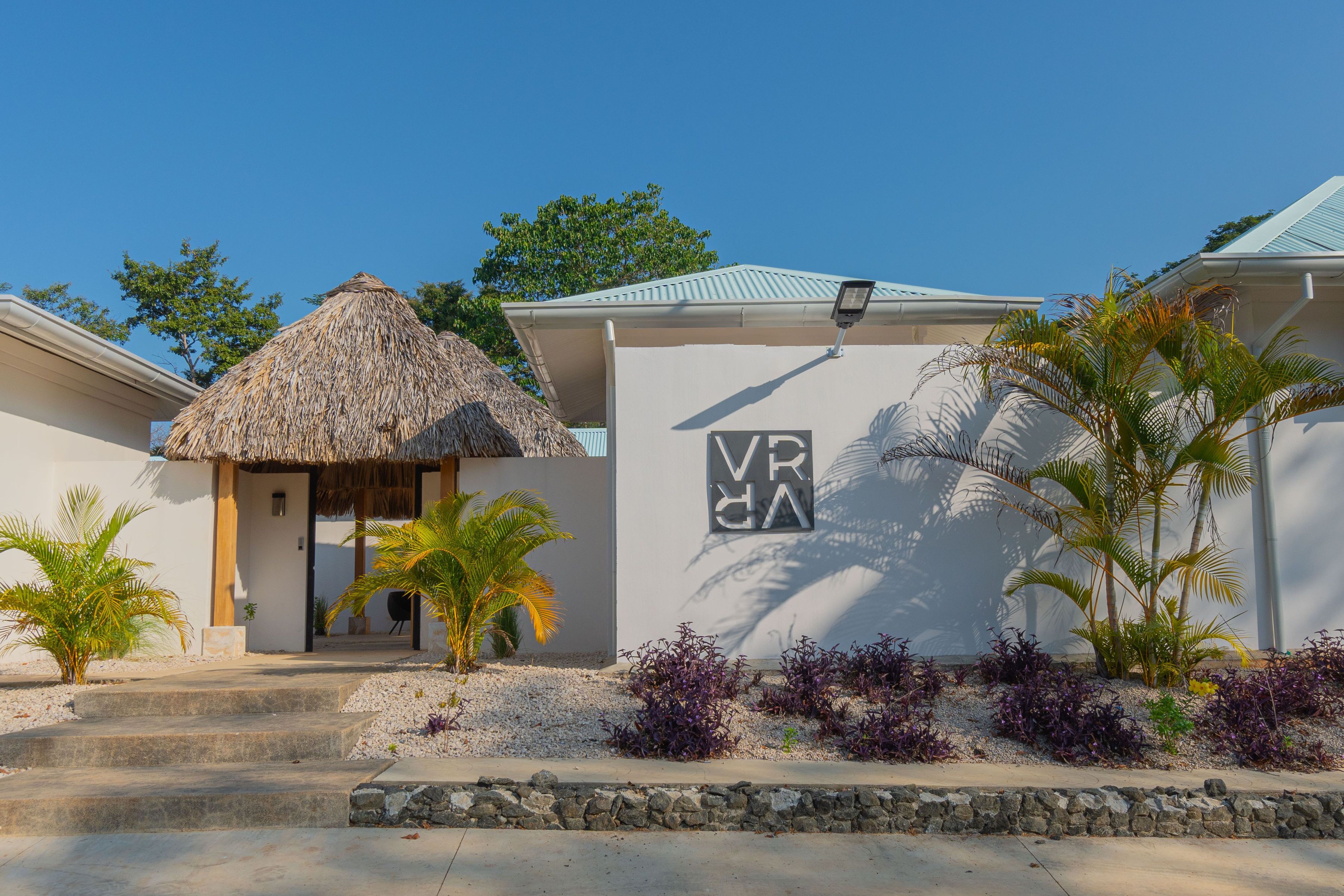 Un bâtiment doté d’une structure à toit de chaume, d’une enseigne indiquant « VR » et « VA », et agrémenté de plantes tropicales.