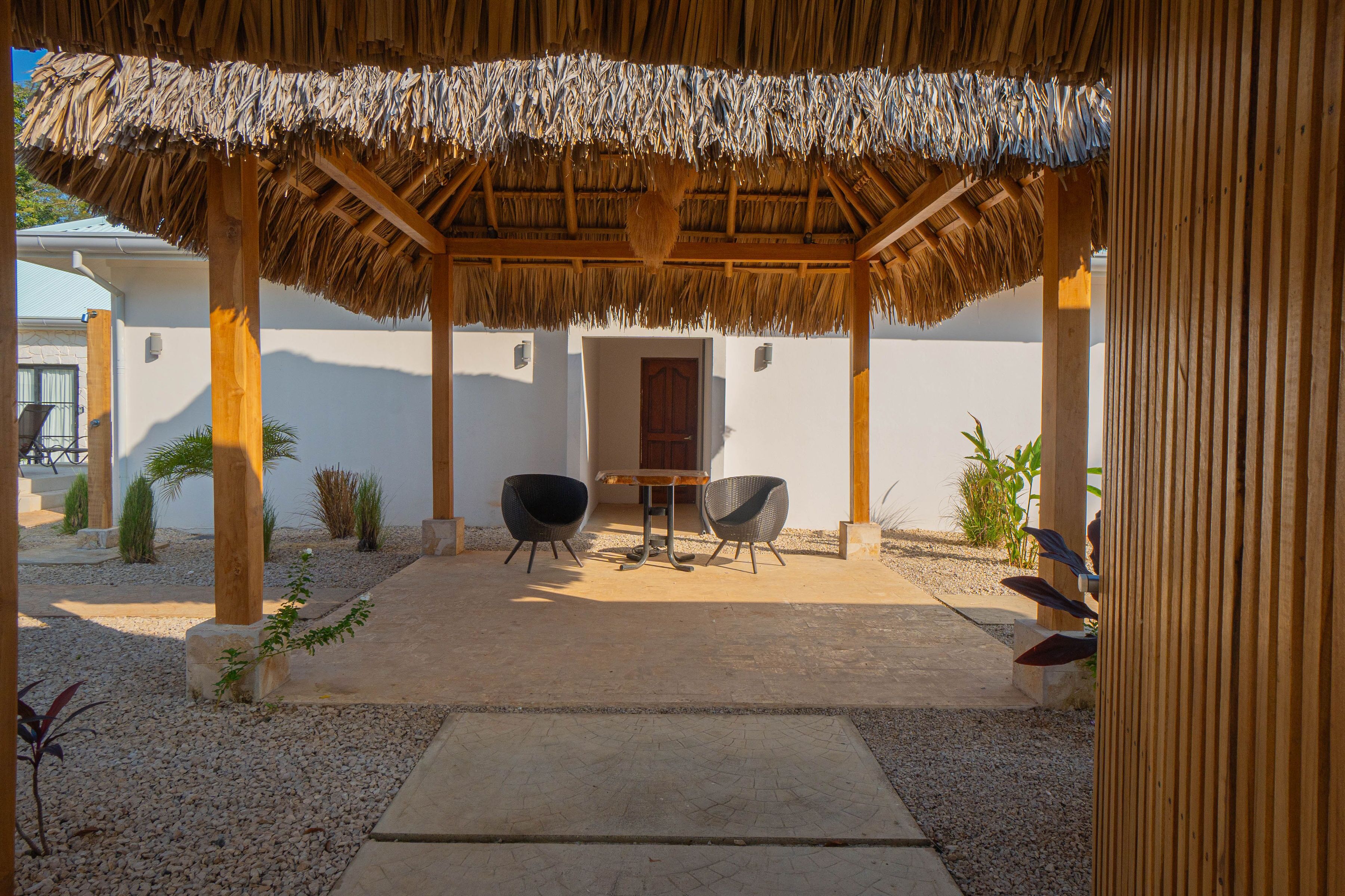 A covered outdoor seating area with two chairs and a table, surrounded by a gravel path and small plants.