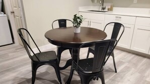 Un coin repas avec une table ronde et quatre chaises, une plante en pot posée sur la table, et une cuisine blanche en arrière-plan.