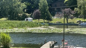 A grassy area with a dock and a pond, surrounded by trees and a building in the background.