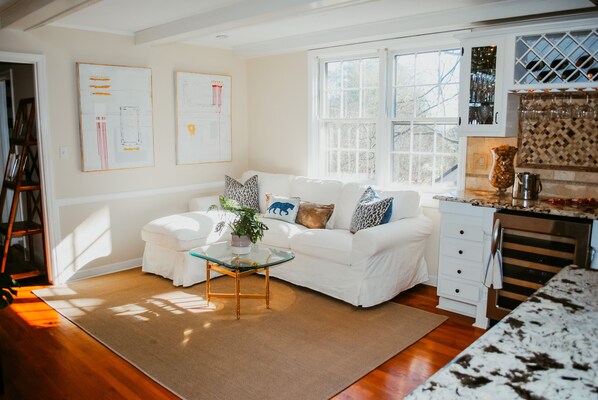A modern living room with a white sofa, glass coffee table, and a kitchen area with white cabinets and a granite countertop. - House on the Hill--The Perfect Master's Rental! (Augusta)