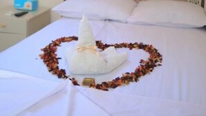 Una cama impecable con un arreglo de flores secas en forma de corazón y un adorno cónico blanco sobre la colcha.
