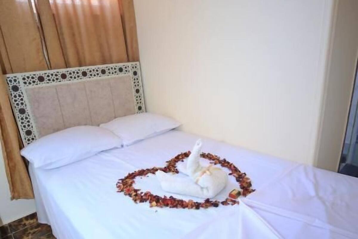 Ein Bett mit weißen Leinentüchern, einem dekorativen Kopfteil und einer Anordnung aus Handtuch und Rosenblütenblättern auf dem Bett.