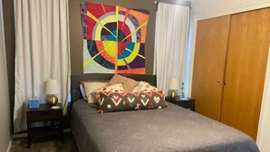 Un dormitorio con cama, un cuadro abstracto de colores, un armario y una mesita de noche con lámpara.