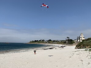 A beach with a clear sky, a person flying a kite, and a building with a clock tower in the background.