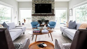 A modern living room with a stone fireplace, two blue armchairs, a grey sofa, and a coffee table with books and remotes.