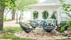 A rustic outdoor seating area with a stone fire pit, surrounded by black wire chairs, under a white building with a tiled roof. - Walkable to Fayetteville + Wine Bar | Sleeps 3 (Fayetteville)