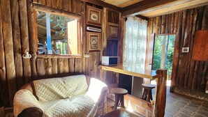 A rustic cabin interior with a stone floor, wooden walls, a wooden ceiling, a sofa, a table, and a window with a view of greenery. - Cabaña Pipil en Los Naranjos (Los Naranjos)