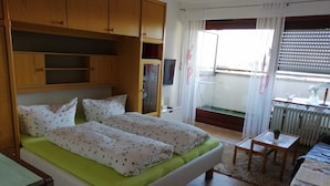 Un dormitorio con cama, armario de madera, una mesita y una ventana con cortinas.
