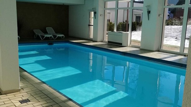 Una piscina interior con agua azul cristalina, suelo de baldosas y dos sillones de descanso.