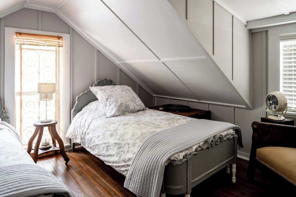 Ein Schlafzimmer mit schrägen Wänden, einem Bett mit weißer Bettdecke, einem Nachttisch mit Lampe und einem Ventilator auf dem Nachttisch.