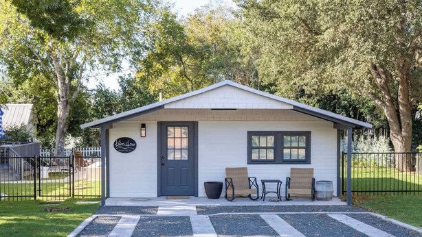 A small, single-story building with a white exterior and a dark roof. It features a front door, two windows, and a small porch area with a bench and a table. - Round Top Cottage for 4 | IN TOWN | Walk to Shops (Round Top)