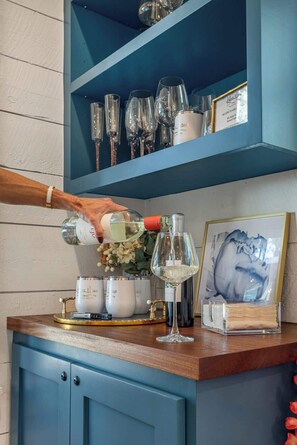 A person pouring a clear liquid into a wine glass on a wooden counter with a blue cabinet and shelves above containing various glassware.