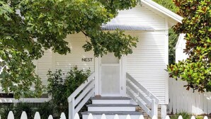 Une maison blanche avec un porche à l’avant, un escalier et une enseigne sur laquelle on peut lire « NEST ».