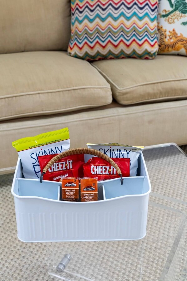 A white plastic basket containing snacks and a bag of Skinny Pop popcorn on a glass table.