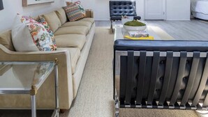 Un salon moderne avec un canapé, une table basse et un pouf noir. Un grand miroir orne le mur, et une porte donne accès à une autre pièce.