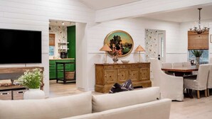A living room with a sofa, a dining table with chairs, a TV, and a wooden cabinet with a painting. - Pool + 2 homes + Sleeps 10 | 6 Min. to Round Top (Round Top)