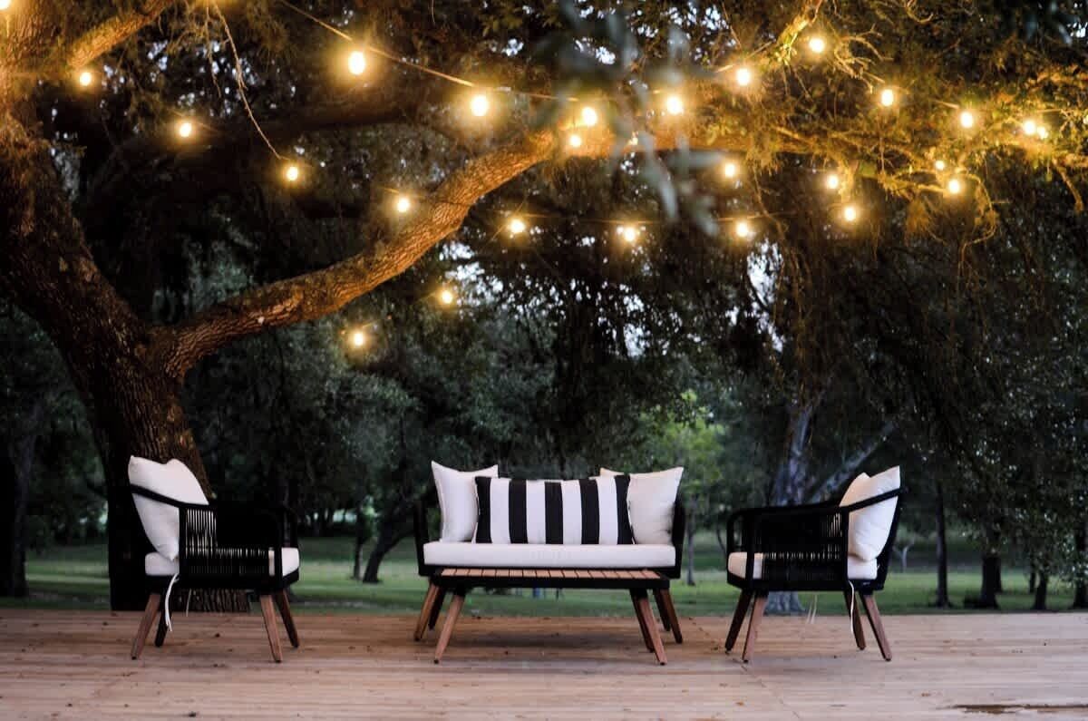 A wooden deck with a white sofa and two black chairs, surrounded by trees and string lights.