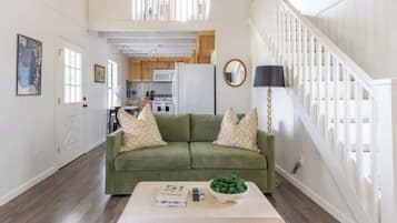 A living room with a green sofa, a coffee table with books and a bowl of green items, a kitchen in the background, and a staircase with white railings.