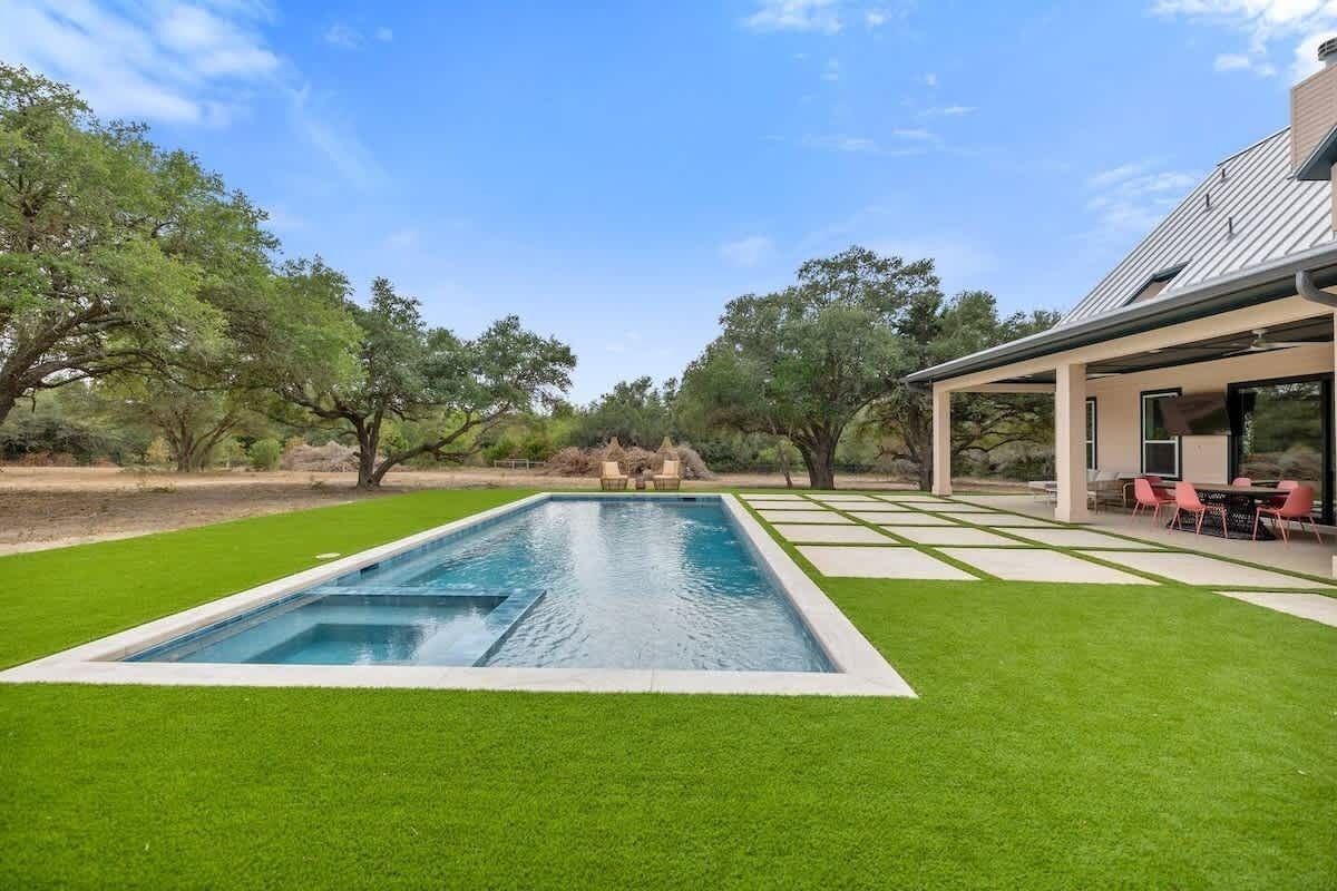 A rectangular swimming pool with a surrounding grassy area, a covered patio with seating, and trees in the background.