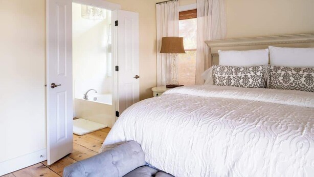 Een slaapkamer met een groot bed, een nachtkastje, een lamp en een badkamer zichtbaar door een geopende deur.