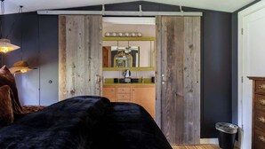 Un dormitorio con una cama negra, un armario de madera y una zona de cocina visible a través de una puerta corrediza.