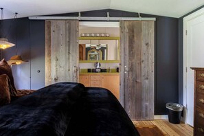 Ein Schlafzimmer mit einem schwarzen Bett, einem Holzschrank und einem durch eine Schiebetür einsehbaren Küchenbereich.