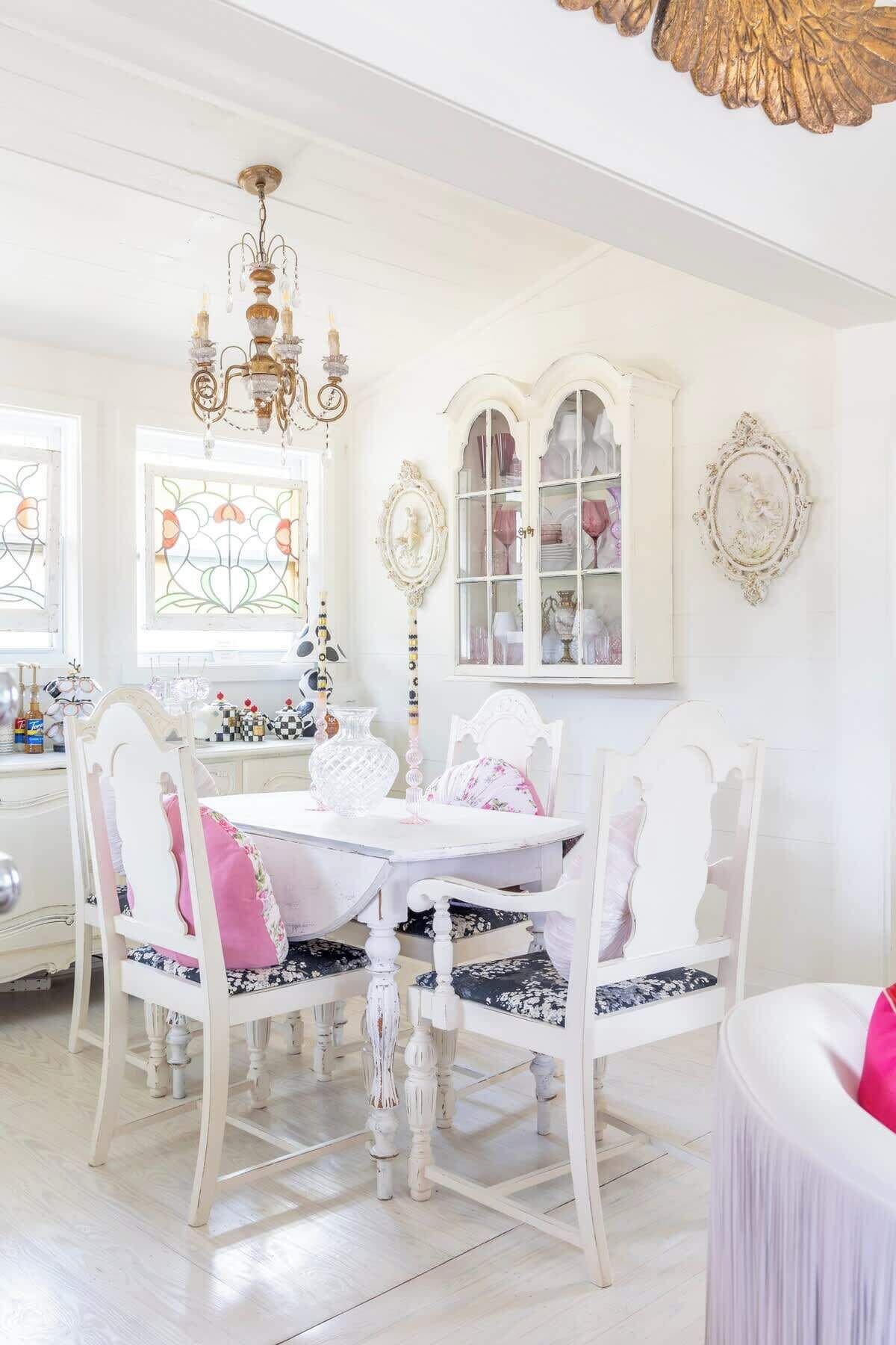 A dining area with a white table, chairs, and a chandelier. There are cabinets with glass doors and a decorative wall piece.
