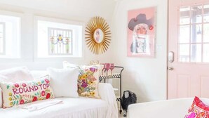 A bright, white room with a bed, a small table with a drink and a book, and a pink door. - Wine, Dine, & Unwind | Stay in Style + Shop RT (Carmine)