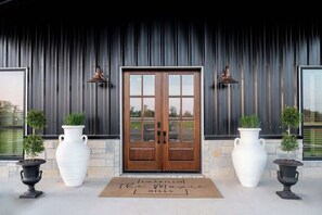 A building entrance with a wooden double door, two large white planters with small trees, and two wall-mounted lanterns. - Casino-style Game Room+Pool+Privacy | The Moxie (Fayetteville)