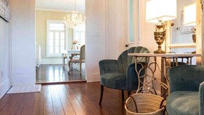 Una habitación con piso de madera, dos sillones tapizados, una silla de mimbre, una mesa con lámpara y vistas a otra habitación con un candelabro.