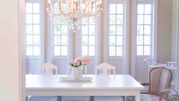 Un comedor con una mesa blanca, dos sillas, una araña y un jarrón de flores.