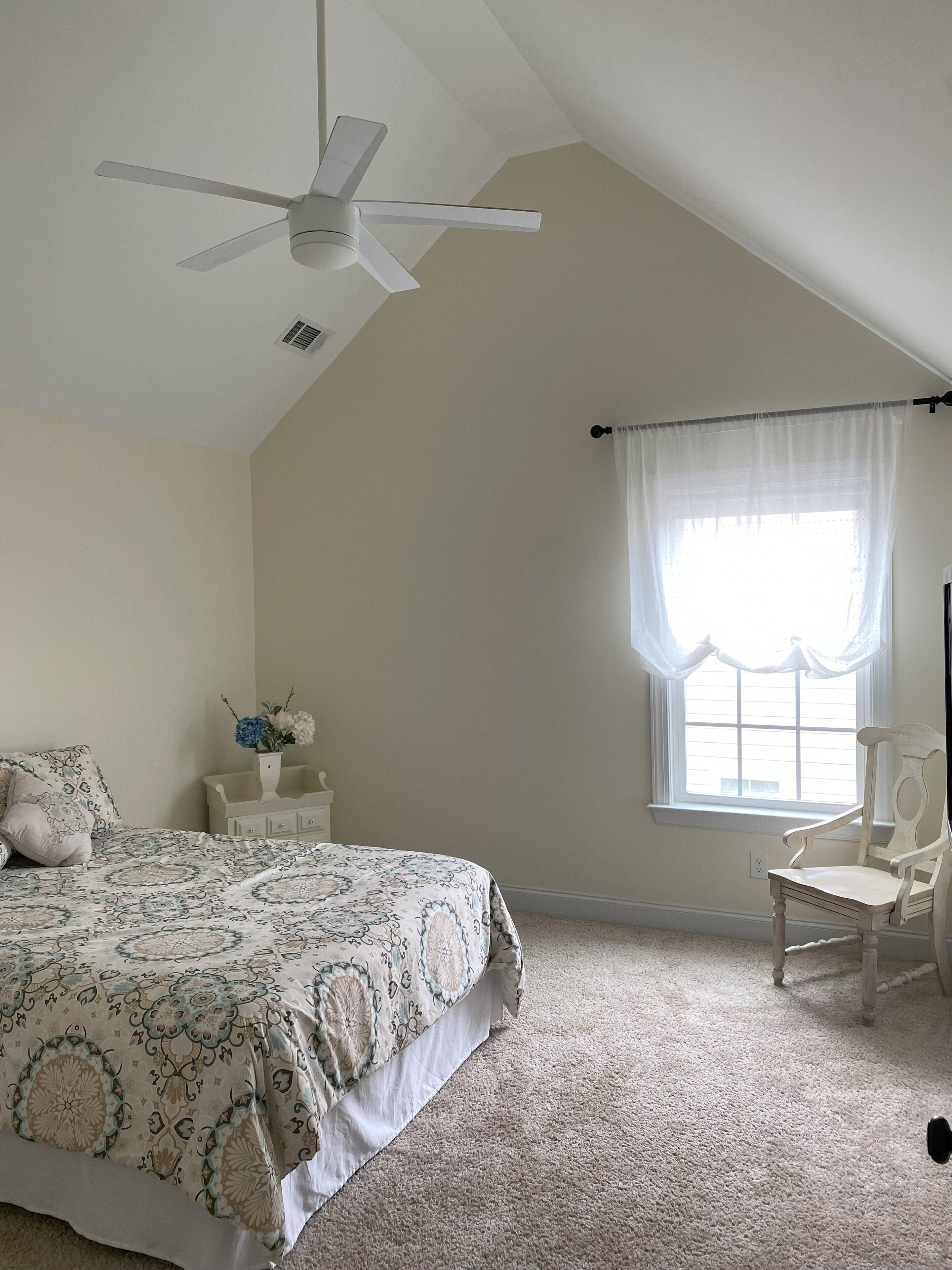 A bedroom with a bed, a chair, a window with curtains, a ceiling fan, and a small table with a vase.